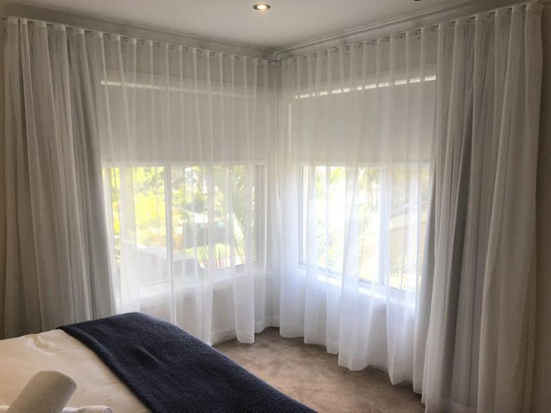 Corner Bedroom Windows With White Sheer Curtains — Winning Blinds in Cardiff, NSW