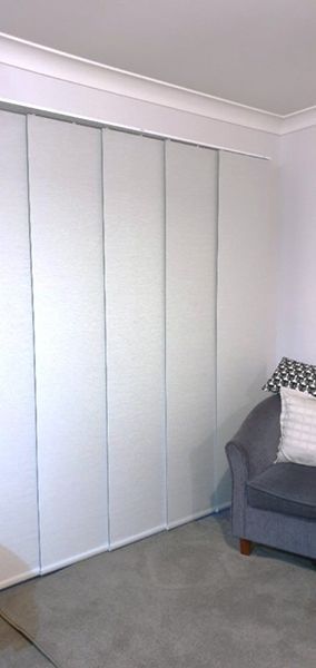 White Panel Blinds Next To A Gray Armchair With Pillows — Winning Blinds in Cardiff, NSW
