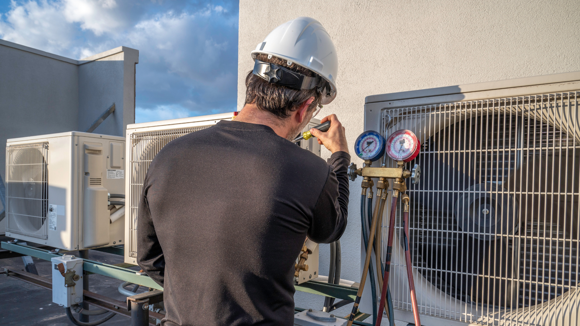 HVAC technician in a hard hat, servicing an outdoor air conditioning unit on a rooftop, using gauges.