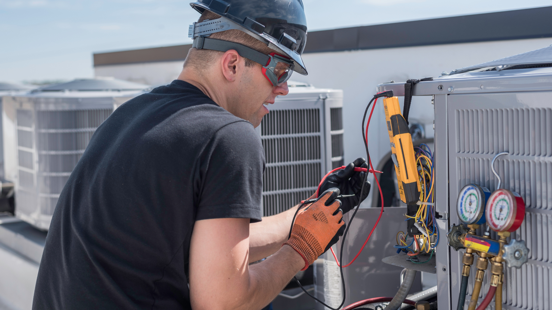 HVAC technician on a rooftop repairs an air conditioning unit. Using a multimeter, gauges, and wearing safety gear.