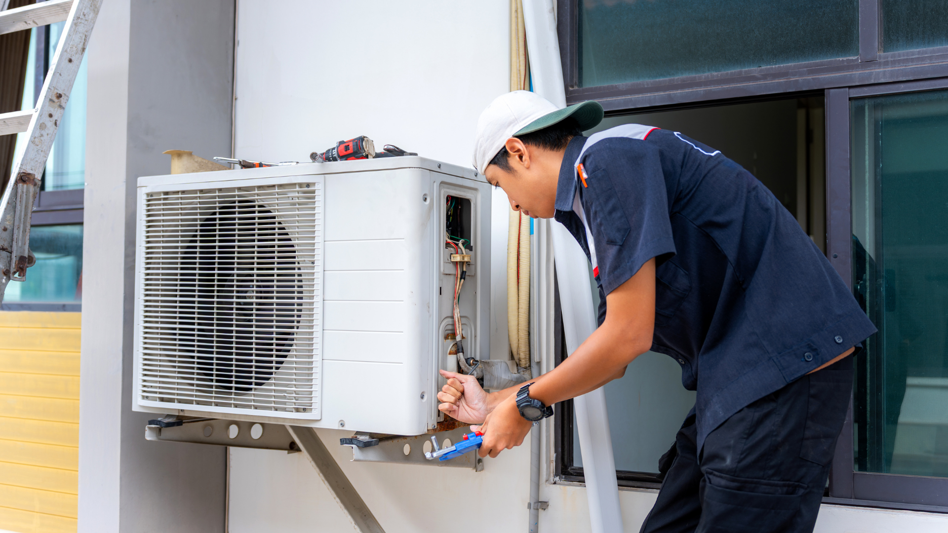 HVAC technician repairs an outdoor AC unit attached to a building, using tools.