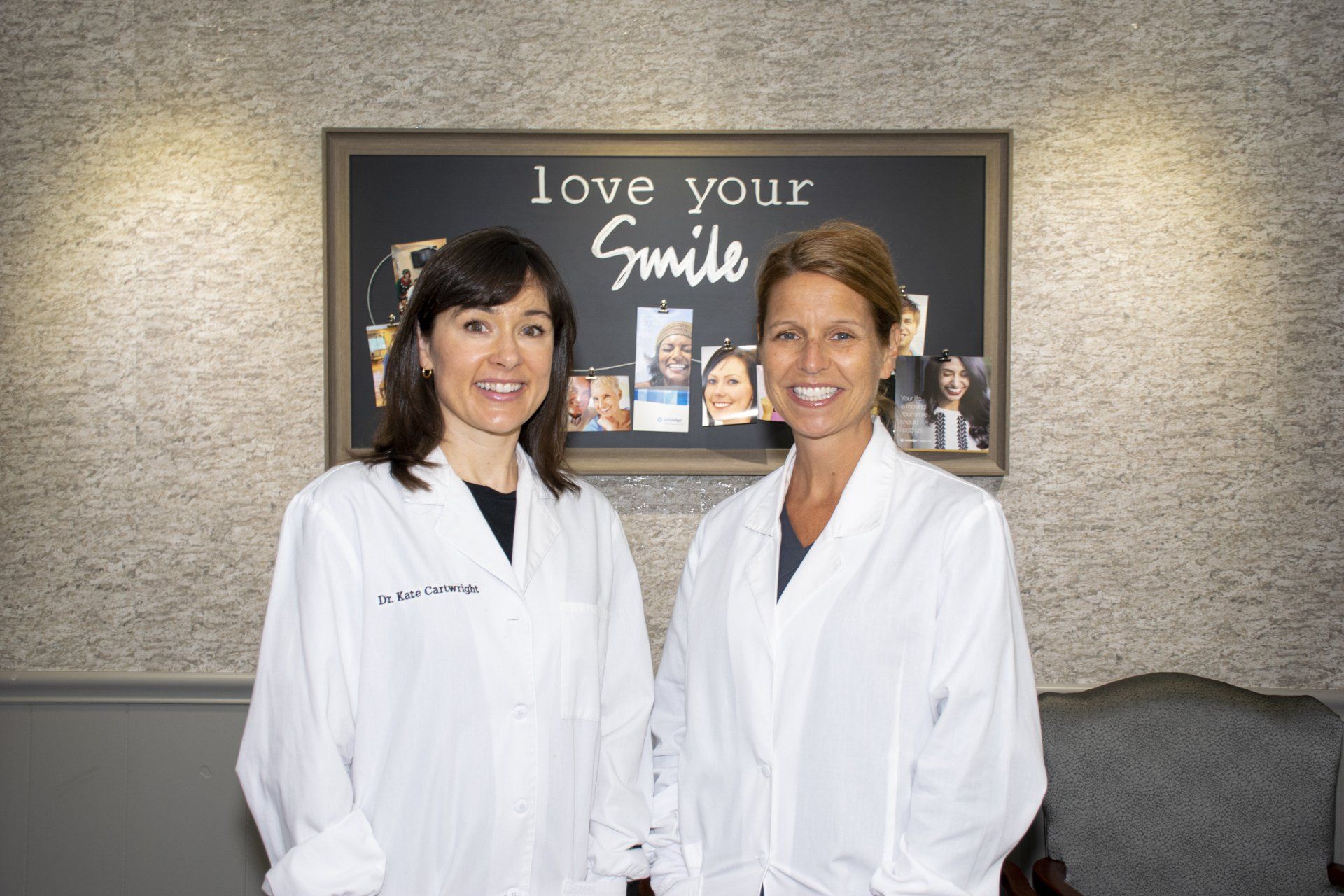 Two women in white coats smiling in a dental office. The sign says 