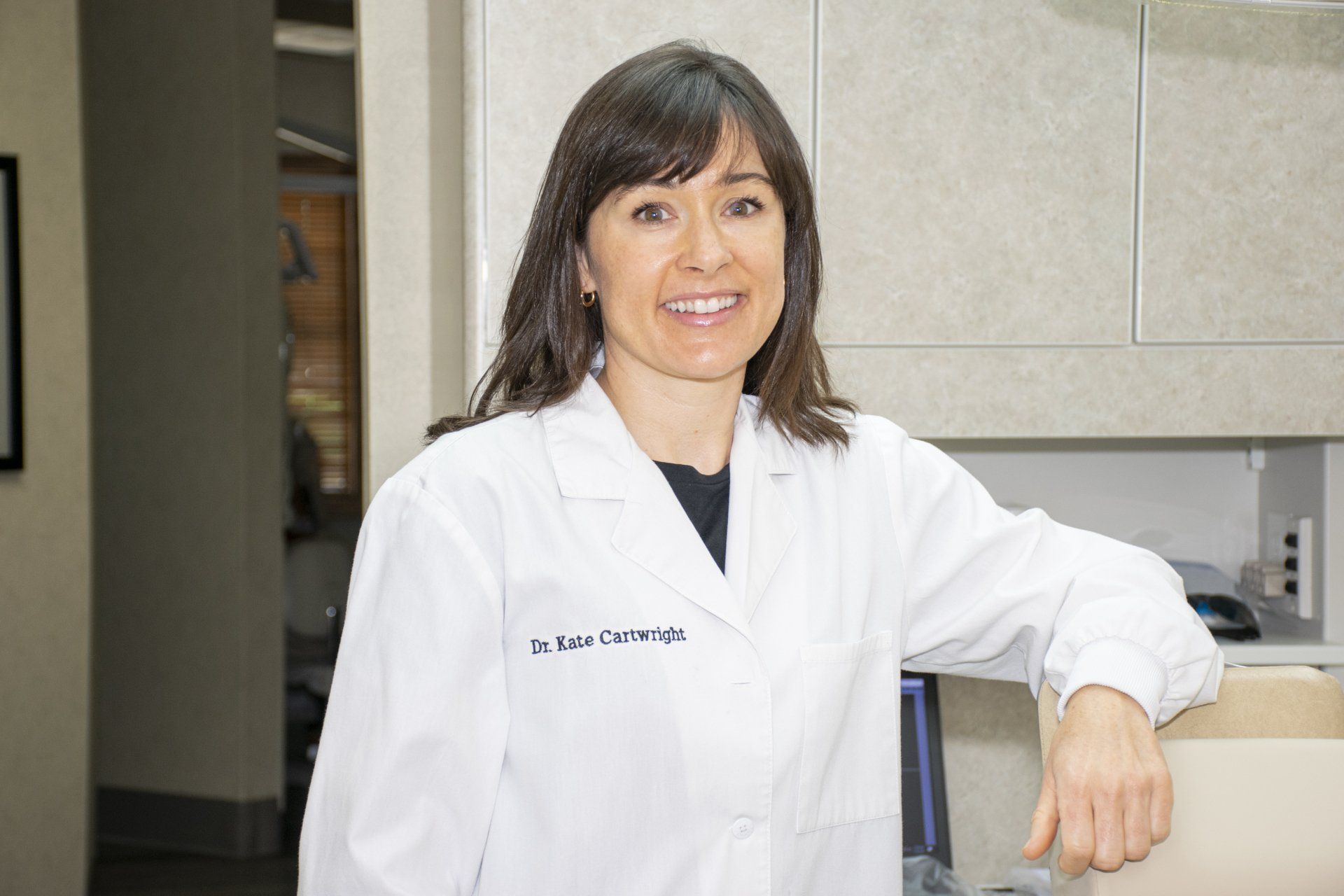 Woman in white lab coat smiles, leaning on a counter in an office.