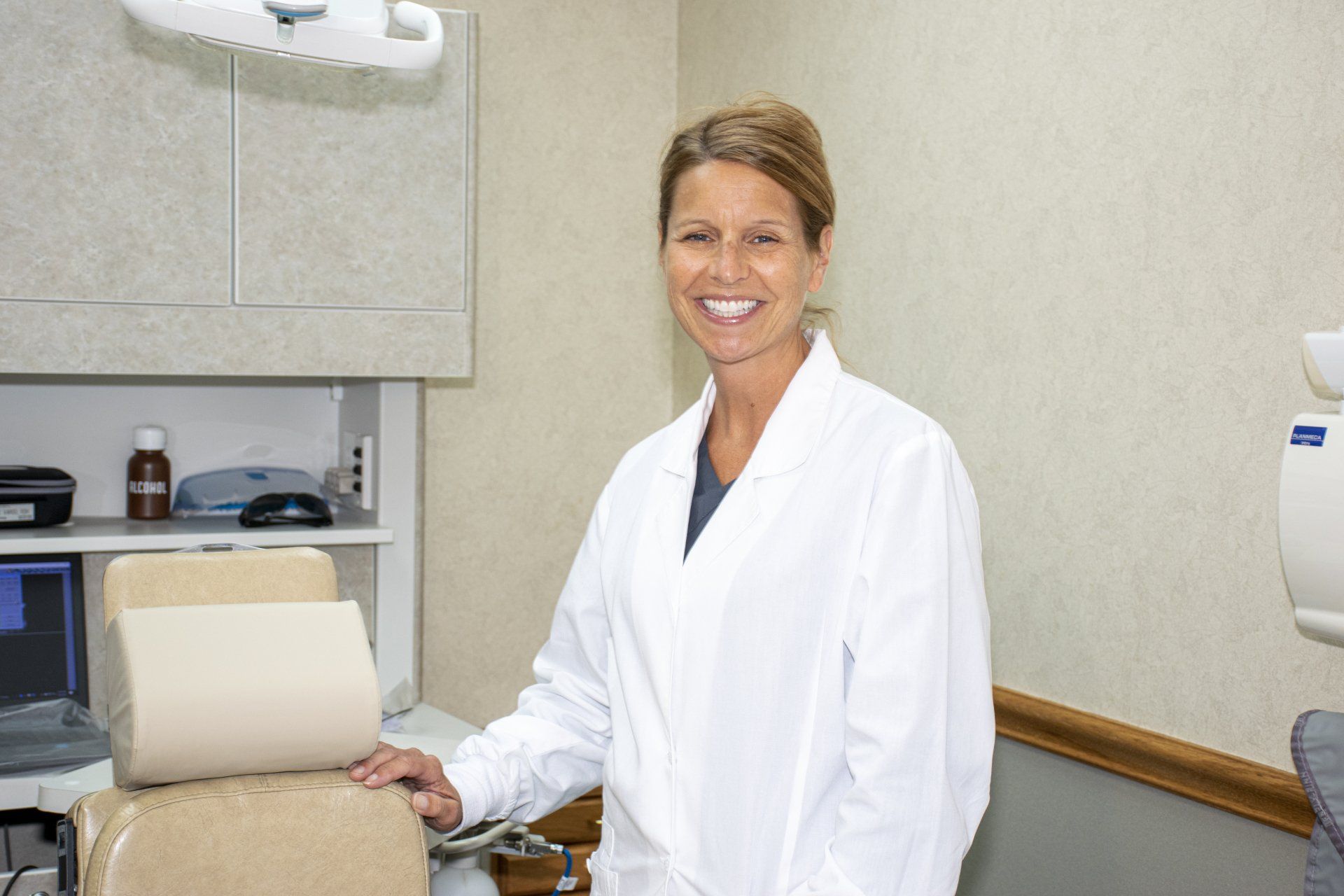 Woman in a white lab coat smiles, standing in a dental office.