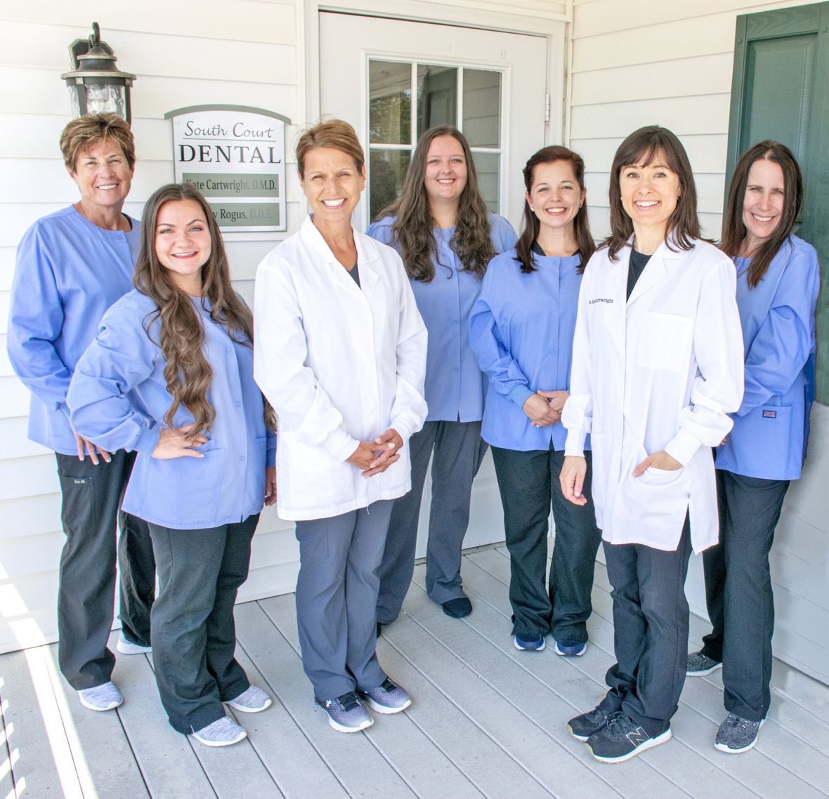 Dental office staff standing on a porch.  They wear scrubs and lab coats.  Smiles are visible.