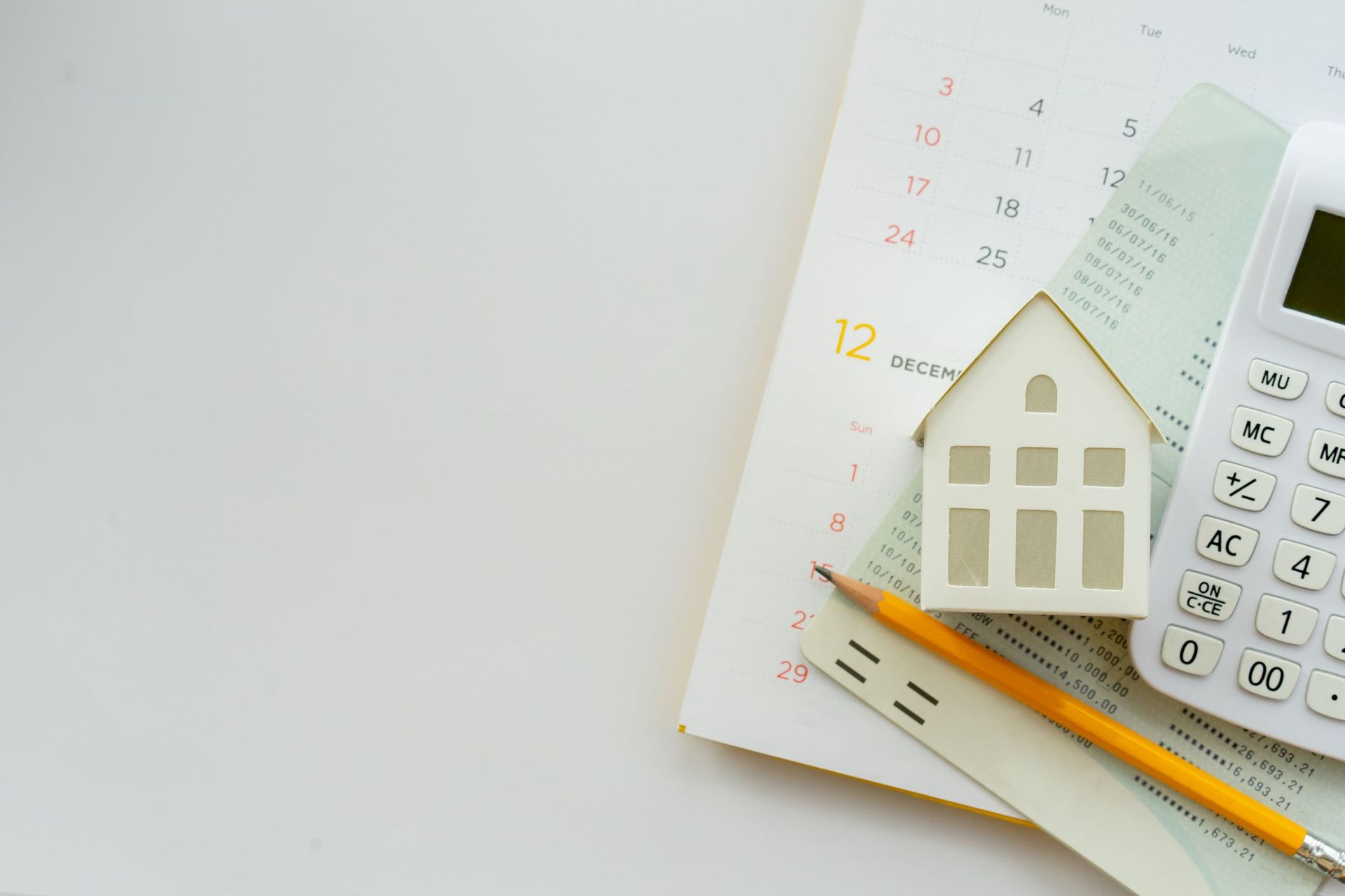 A small house is sitting on top of a calendar next to a calculator and a pencil.