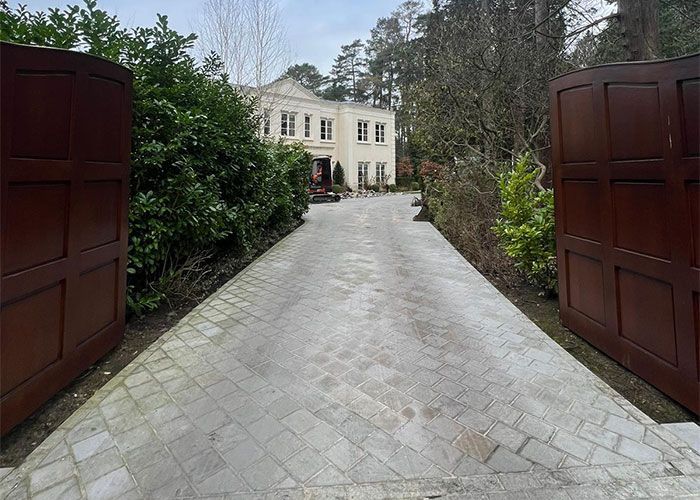 Grand entrance to a Woking estate, showcasing a light grey block paving driveway and dark wooden gates by Surrey Hills.