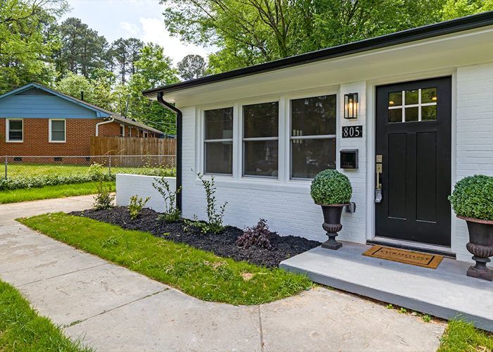 A white house with a black door and a brick house in the background.
