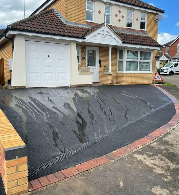 New black tarmac driveway with a decorative brick border in front of a house in Woking by Surrey Hills.
