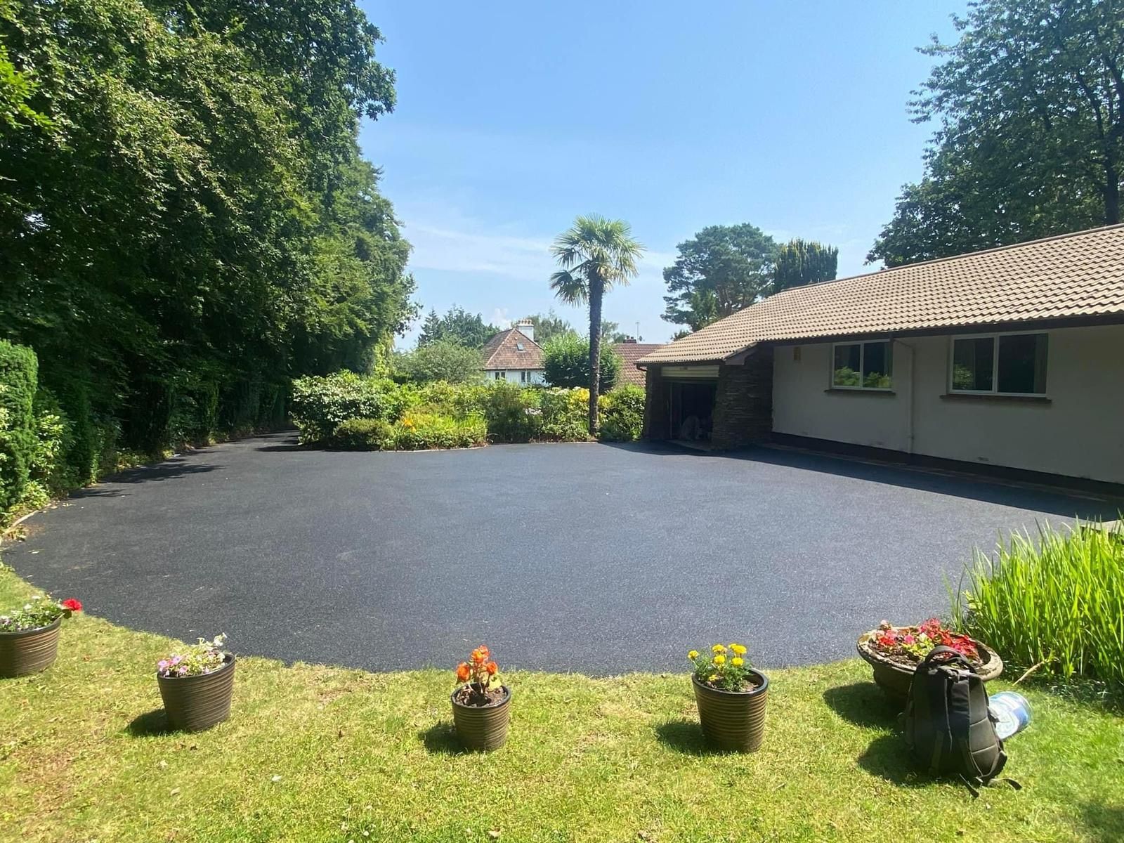 Expansive new tarmac driveway by Surrey Hills Driveways, set amidst lush Woking landscaping with trees and potted plants.