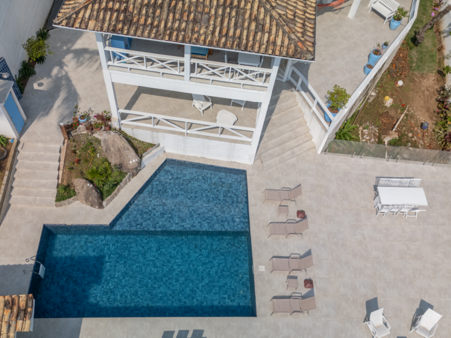 Vista aérea de uma casa com piscina azul, espreguiçadeiras e um pátio com azulejos ao lado de canteiros de jardim.