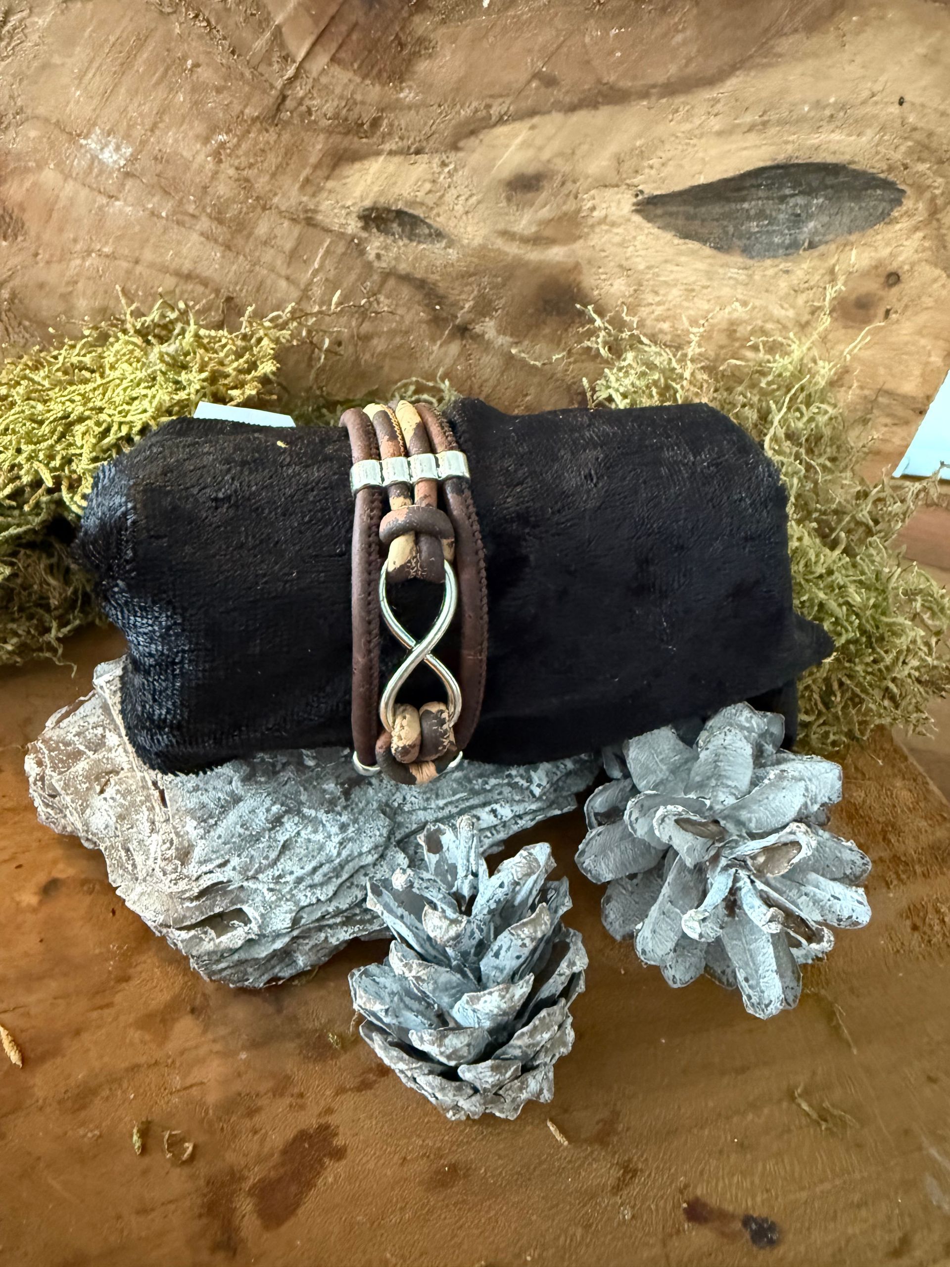 Bruine leren armband met een zilveren oneindigheidssymbool op een zwarte boomstam, versierd met dennenappels.