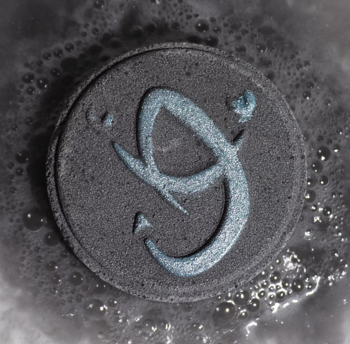 Donkere, ronde zeep of tablet met een lichtblauw abstract symbool, omringd door zeepsop