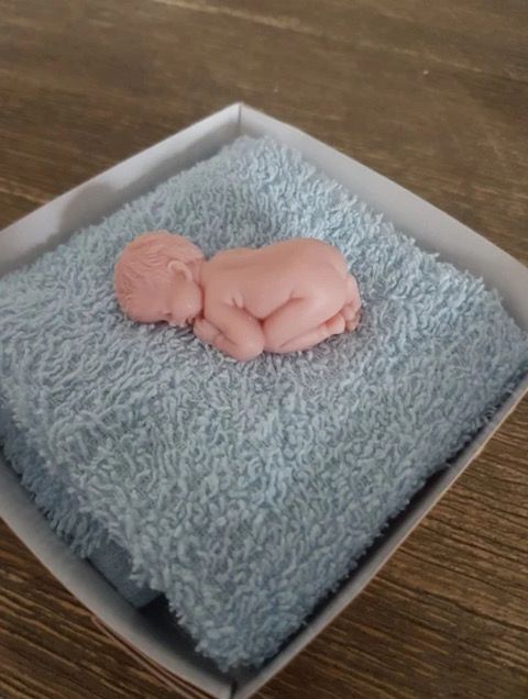 Een babyfiguurtje ligt opgerold op een roze handdoek in een witte doos.