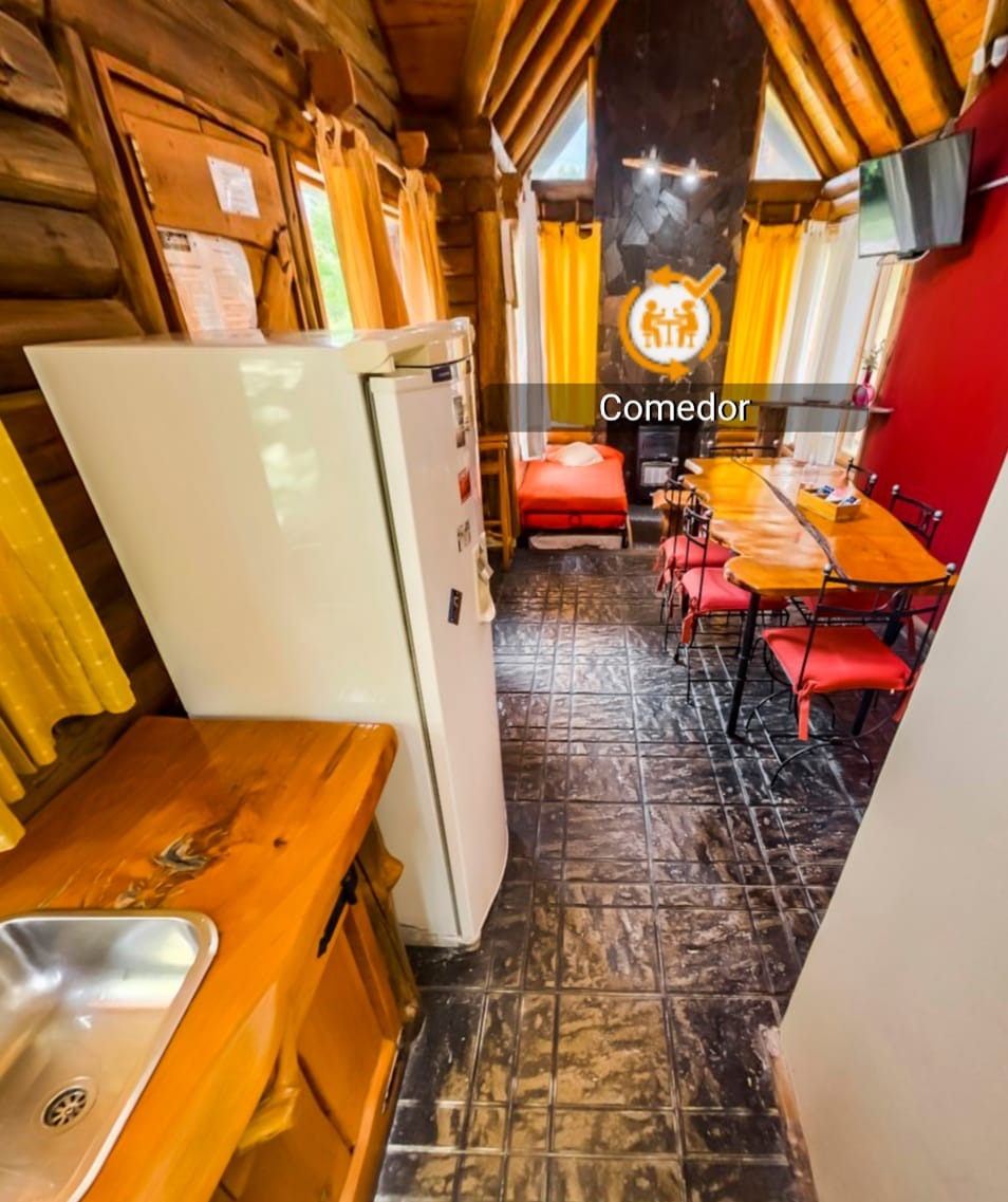 Estrecha zona de cocina-comedor con nevera blanca, armarios de madera, sillas rojas y suelo de baldosas.
