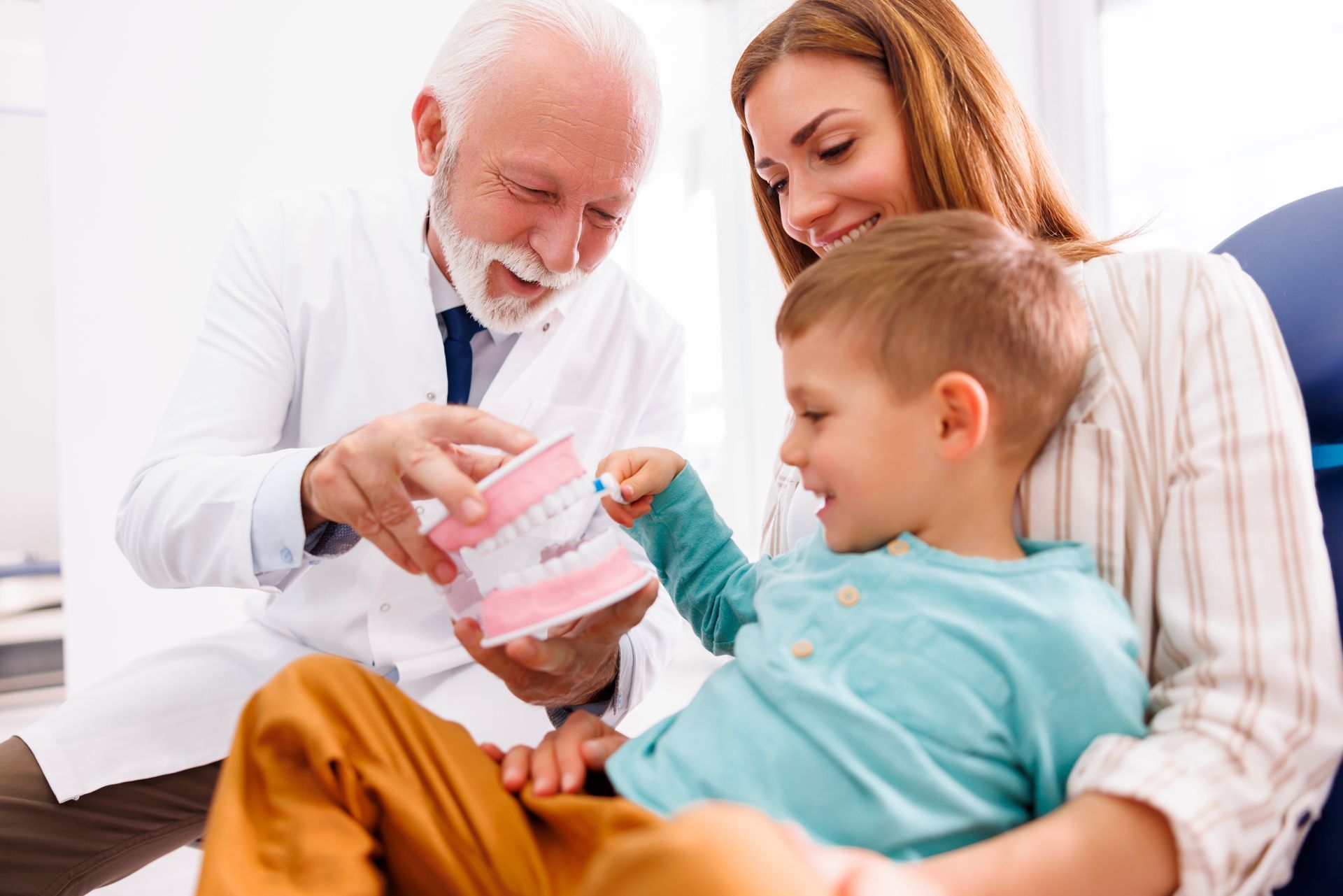 A male dentist is educating a child patient sitting in his mother's lap using a plastic tooth model.