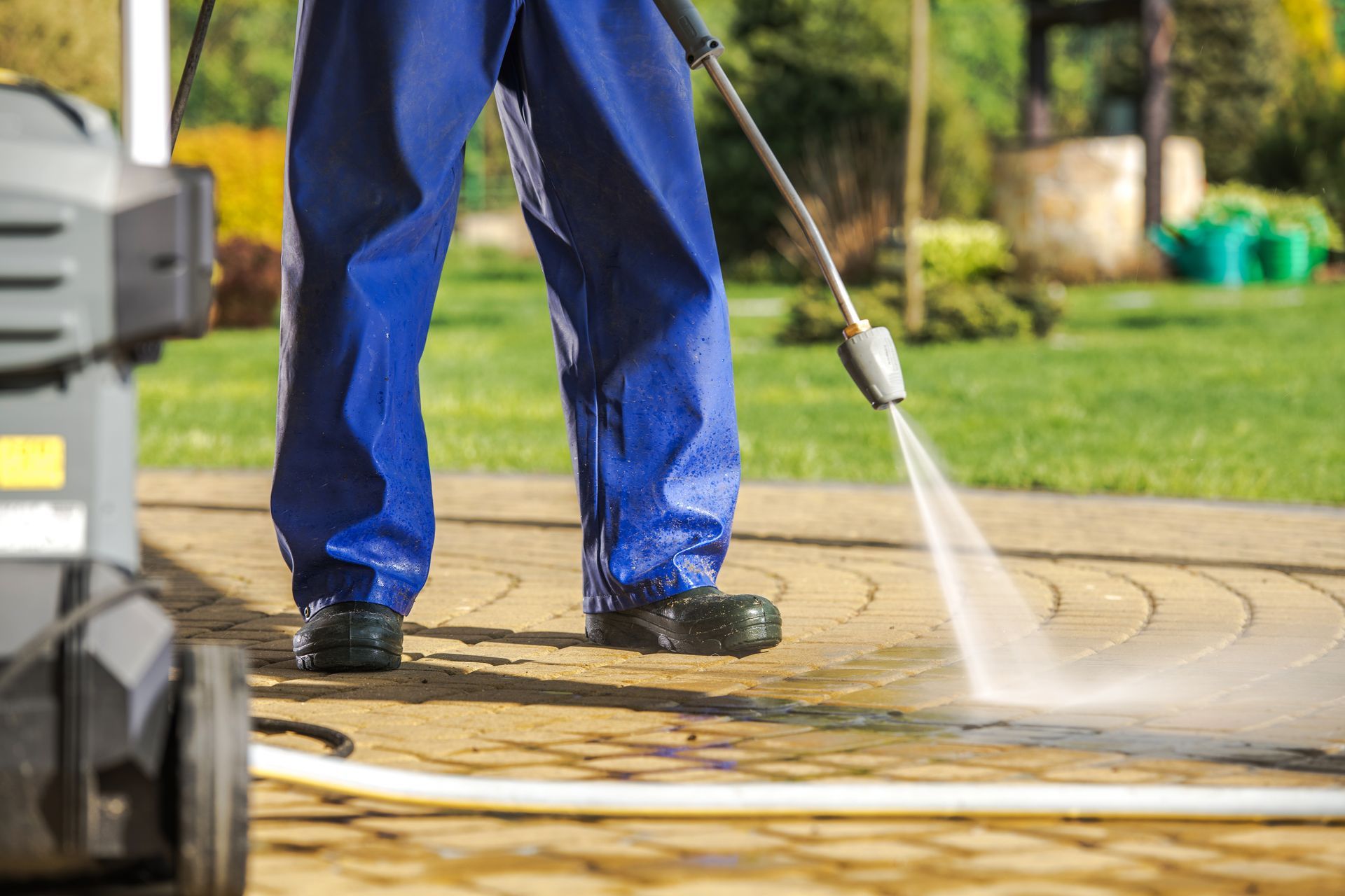 Person in blue overalls power washing a brick patio in a yard.