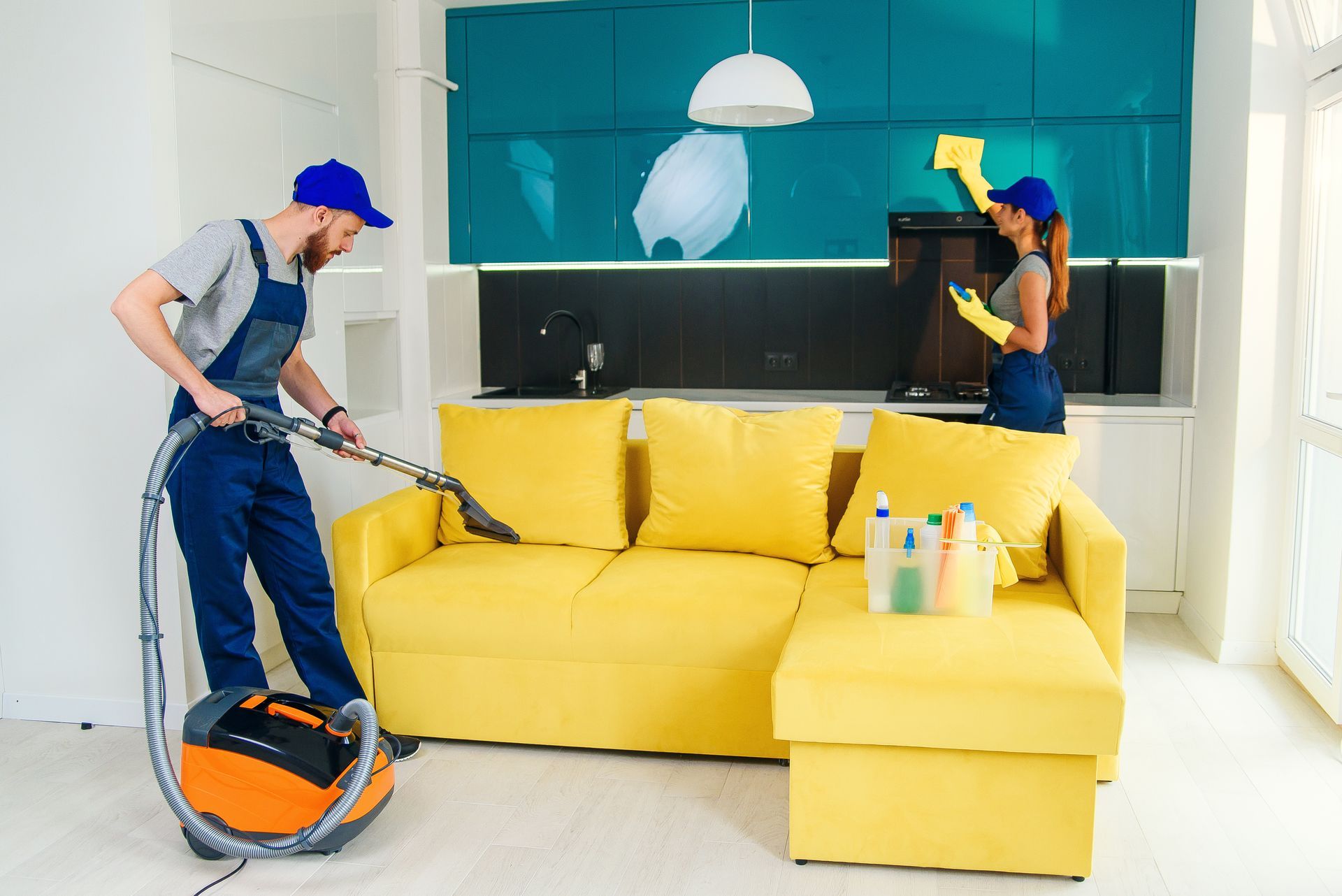 Two people cleaning a yellow sofa in a brightly lit room. One vacuums while the other dusts.