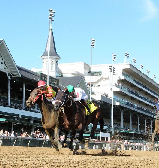 Three jockeys race horses around a dirt track beside grandstands under a clear blue sky.