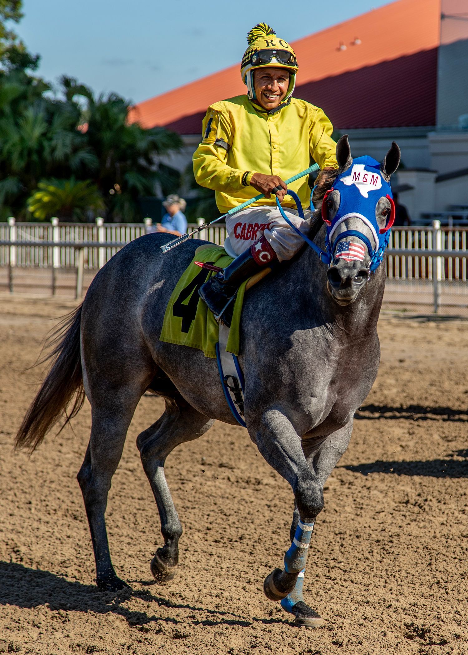 Jinete vestido de amarillo montando un caballo de carreras gris en una pista de tierra, con una máscara facial azul y gradas con techo naranja detrás.