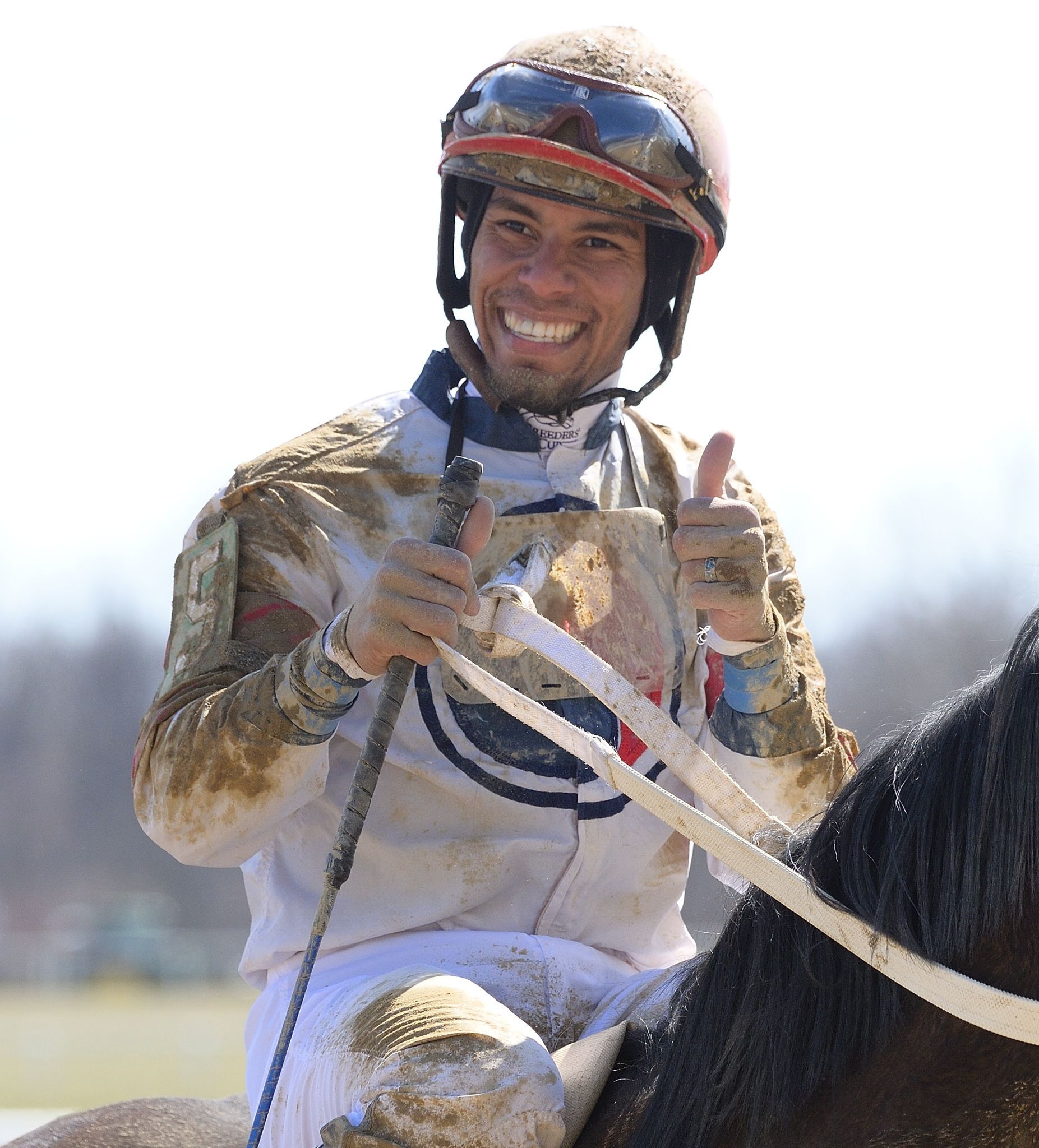 Mud-covered jockey Jamie Rodriguez wearing a helmet and goggles smiles and gives a thumbs-up while sitting on a horse.