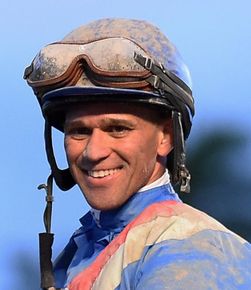 Smiling jockey in blue silks and goggles on a horse, outdoors under a clear sky