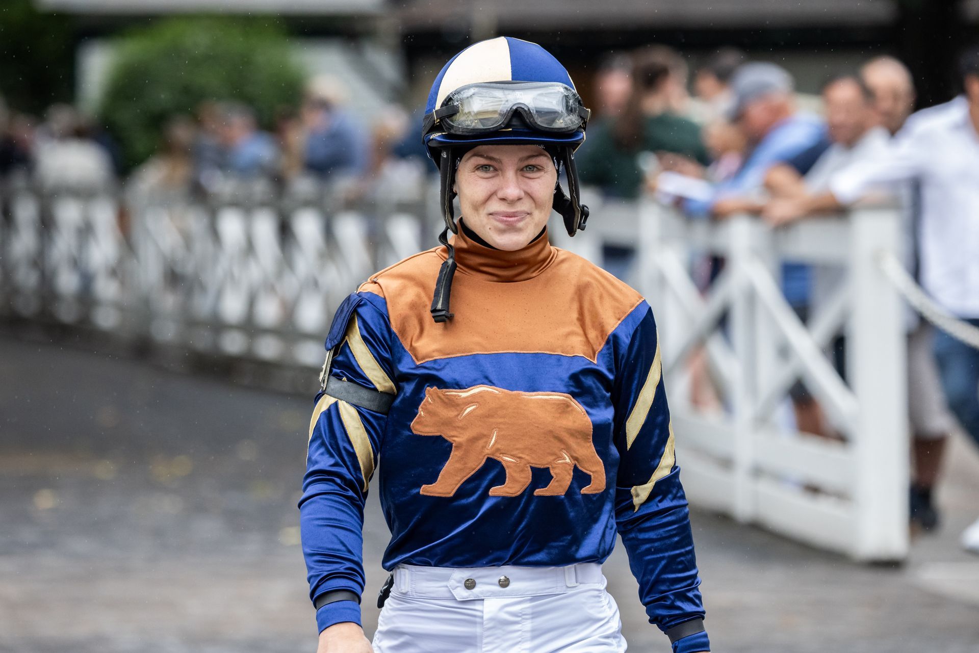Jockey Katie Davis in a blue and copper-colored uniform with a bear emblem on the chest walks past a white fence.