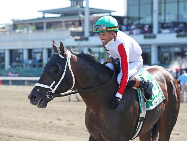 Jockey in green silks riding a brown horse during a race, with spectators blurred in the background