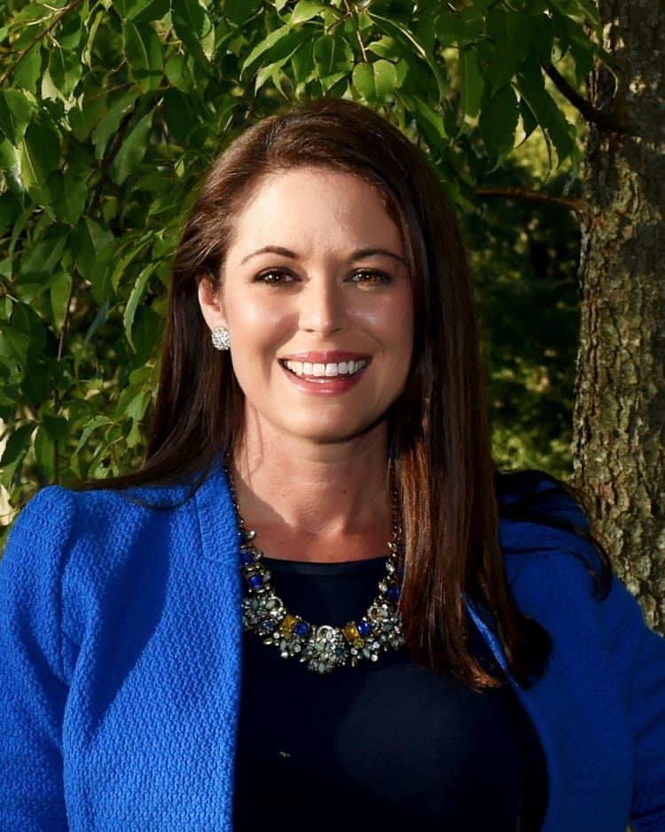 Smiling woman in a blue jacket standing outdoors in front of green foliage