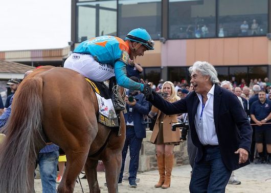 Jockey on brown horse at racetrack, wearing white silks and blue saddlecloth, with scoreboard in background