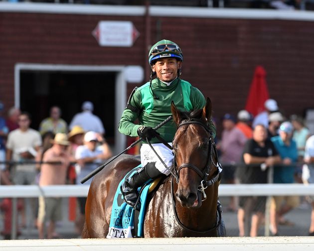 Jockey in green silks riding a brown horse during a race, with spectators blurred in the background