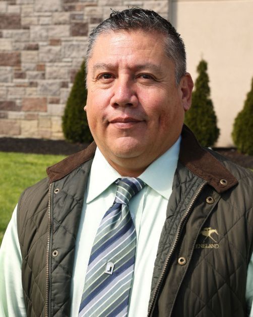 Man in a quilted vest and striped tie standing outdoors in front of a stone wall and greenery