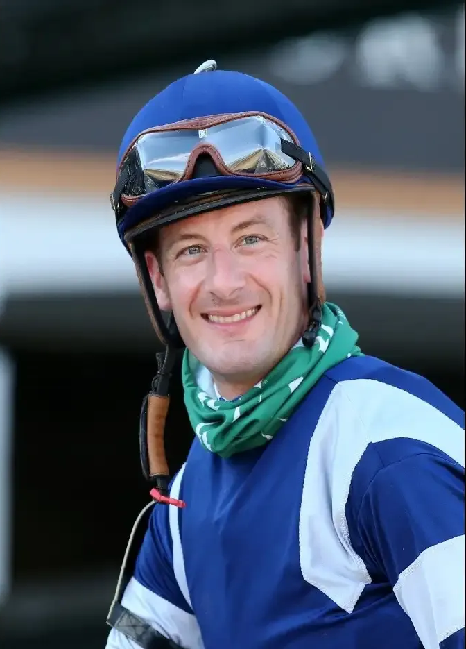 A smiling jockey wearing a blue helmet and racing silks, with protective goggles resting on the helmet brim.