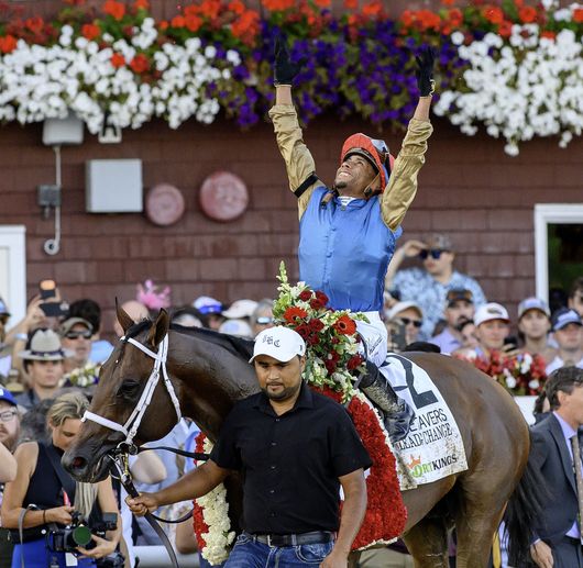 Jockey on brown horse at racetrack, wearing white silks and blue saddlecloth, with scoreboard in background