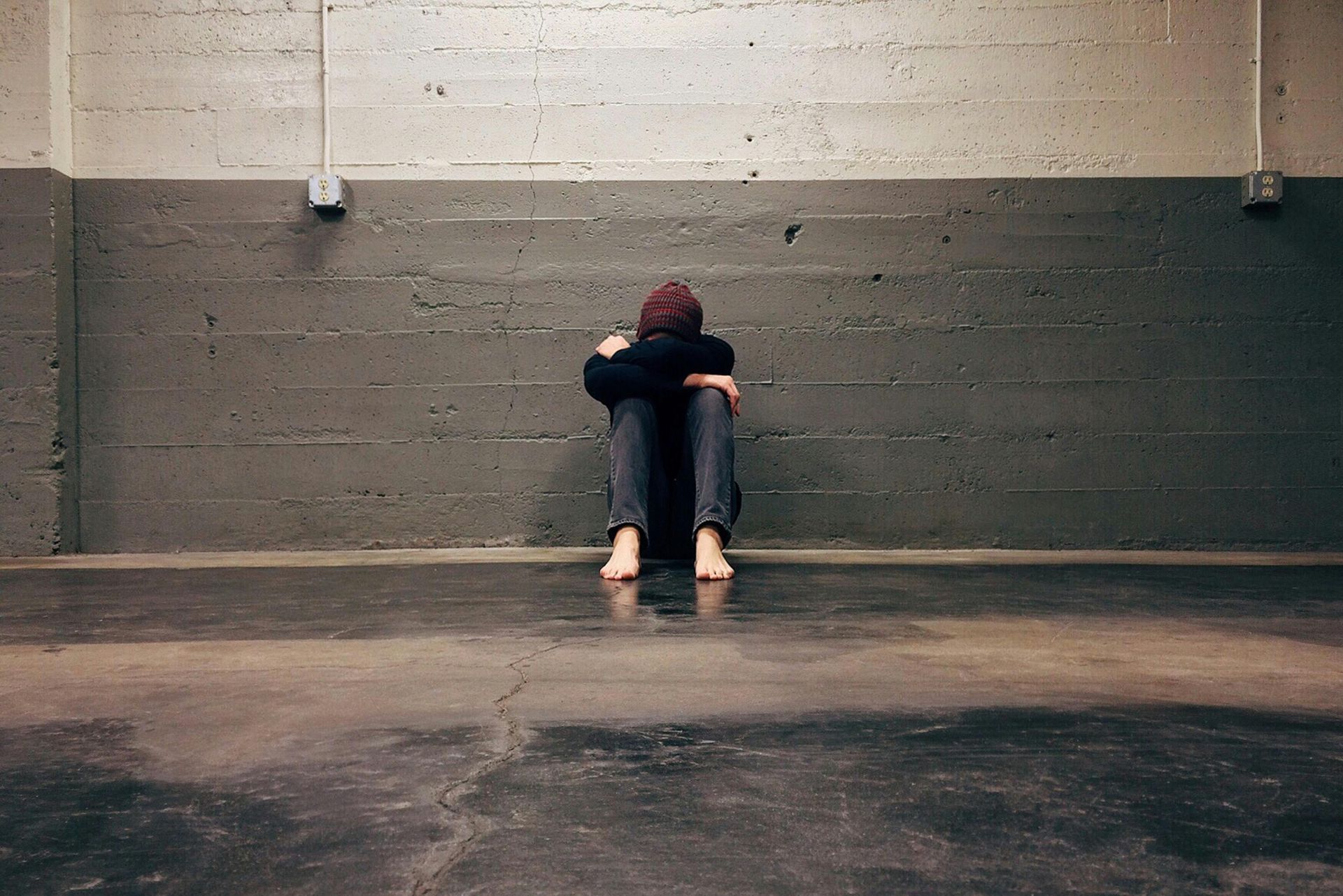 A person sits on the floor, curled up against a gray concrete wall in a large, dimly lit room.