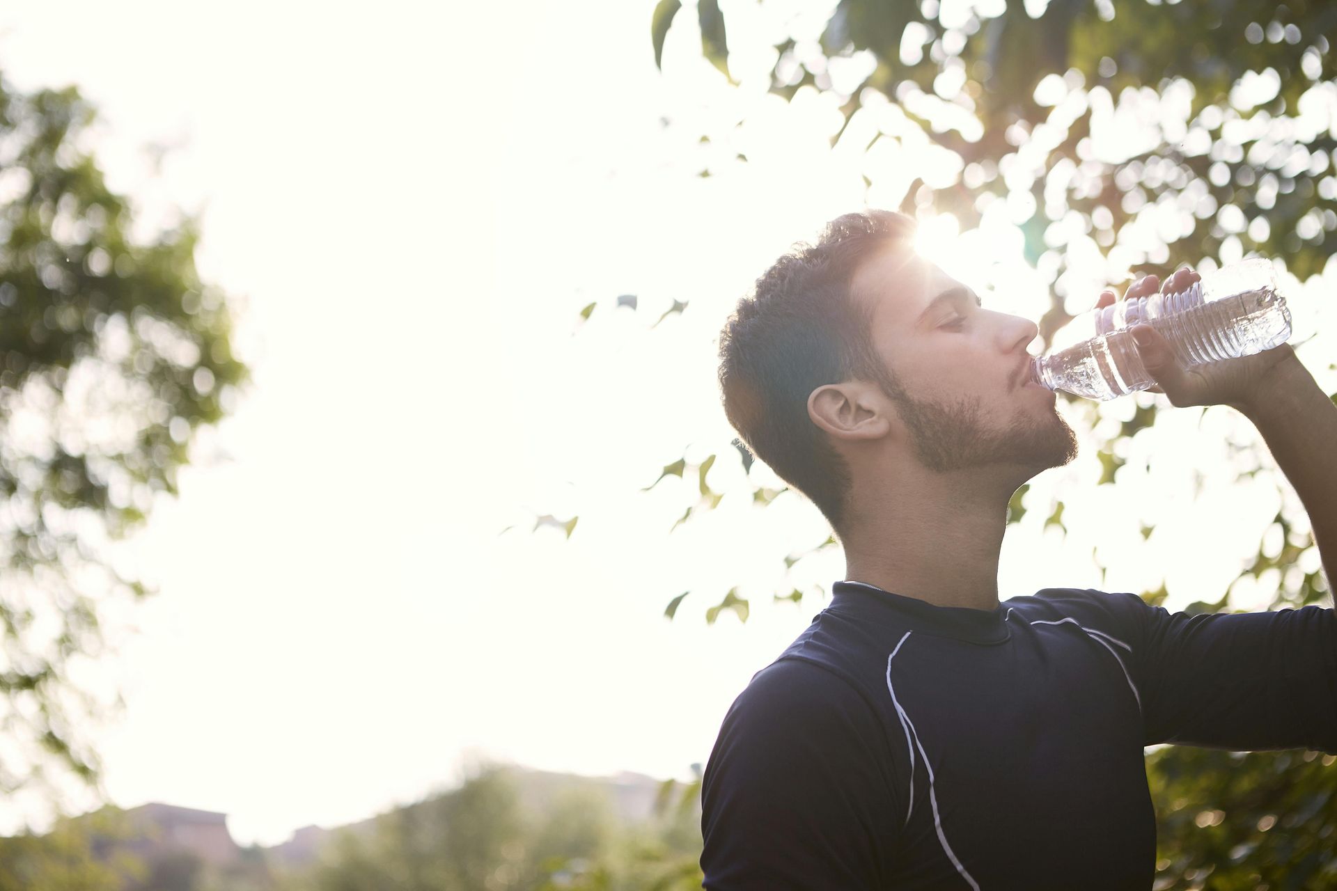 A person wearing a dark athletic top drinks from a clear water bottle outdoors against a bright, sunlit background.