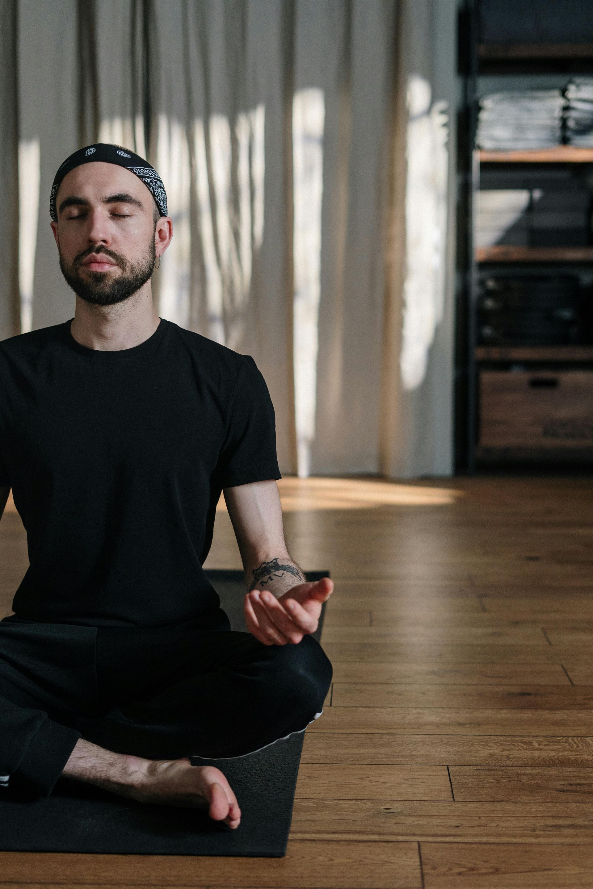 A person wearing a black t-shirt and headwrap sits in a cross-legged meditative pose on a yoga mat in a sunlit room.