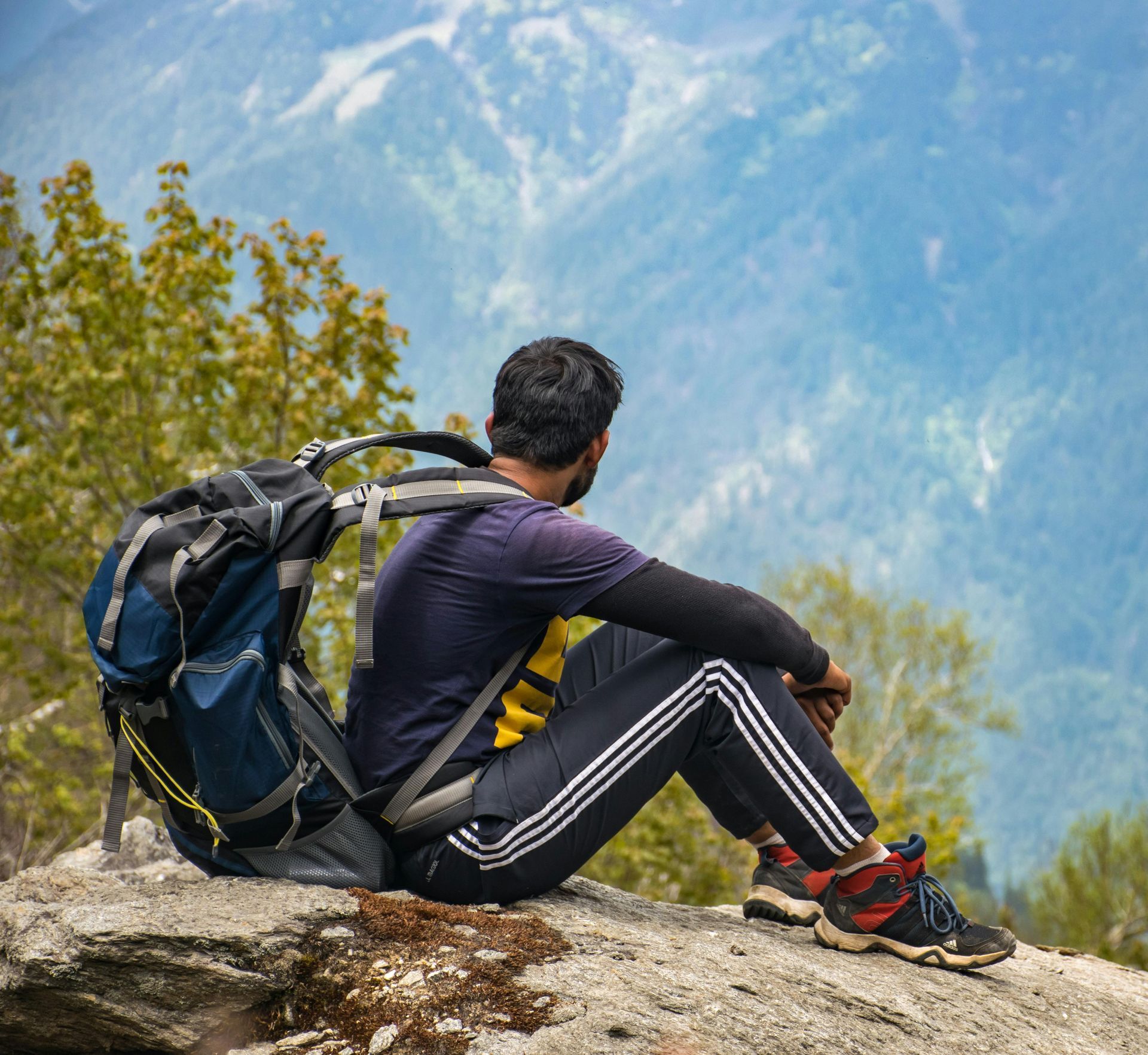 A hiker with a large backpack sits on a rocky cliff, looking out over a vast mountain valley.