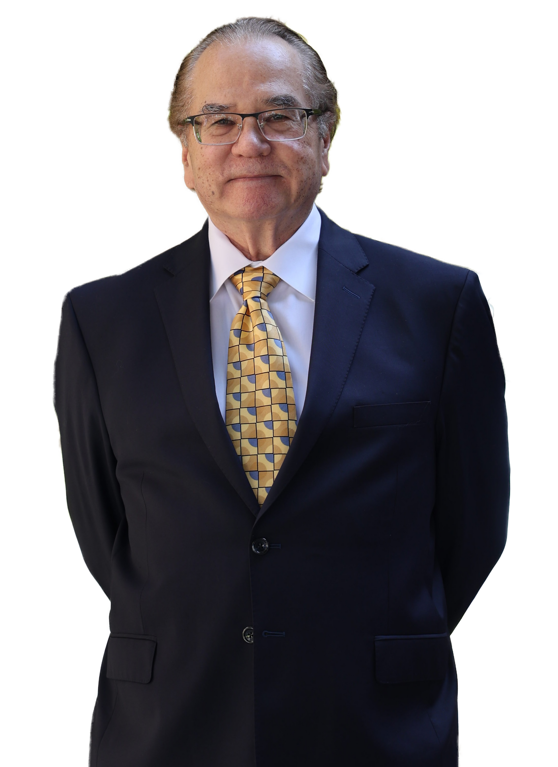 Man in dark suit, glasses, and yellow patterned tie, standing with hands behind his back.