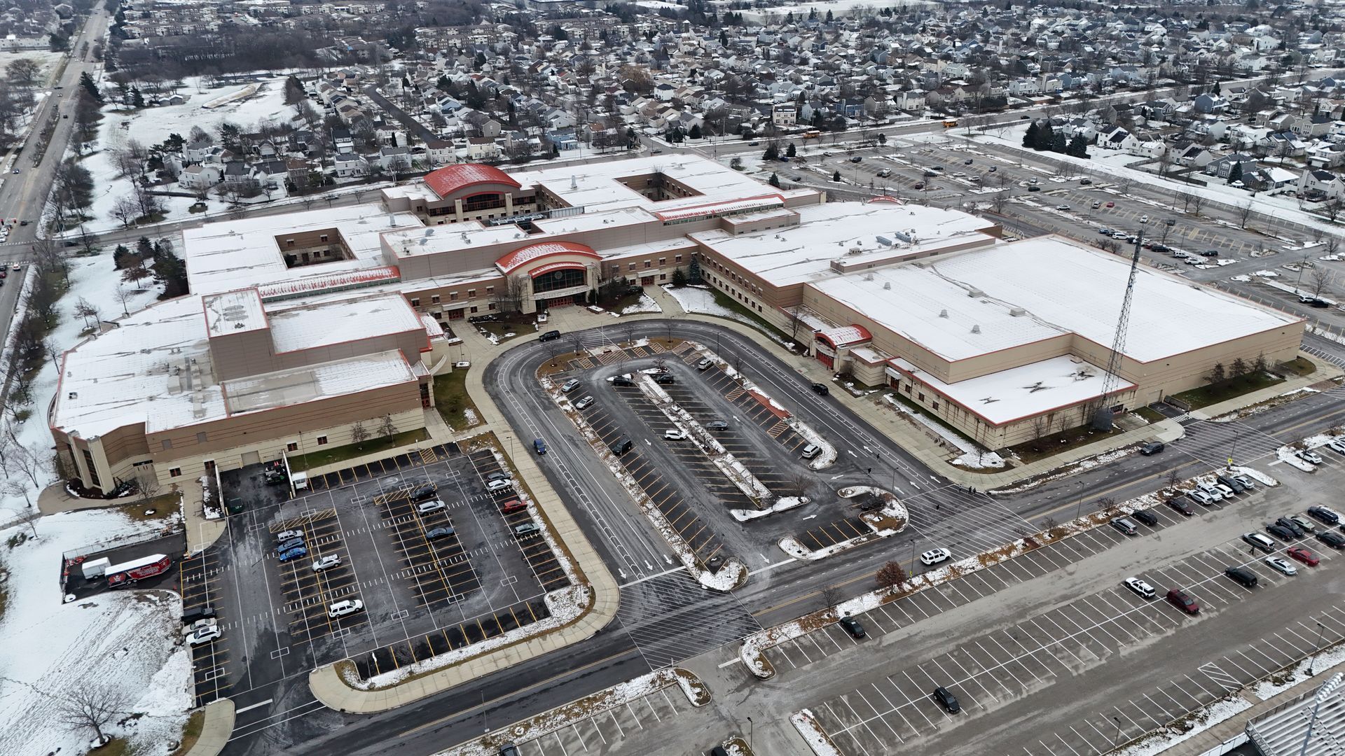 Aerial view of a snowy shopping mall and parking lot, with cars, trees, and buildings visible.