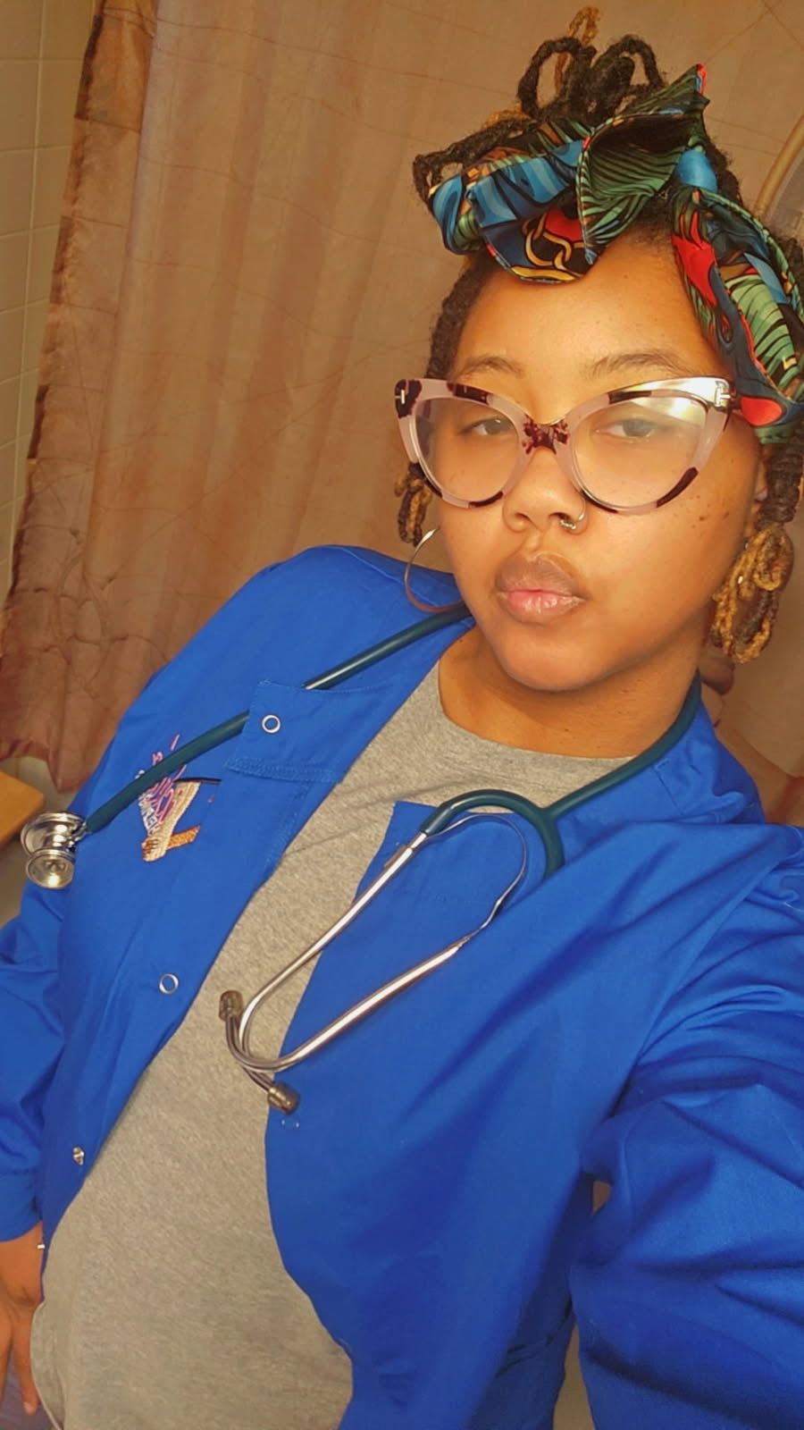 Person wearing eyeglasses, stethoscope, and blue medical scrubs, headwrap.