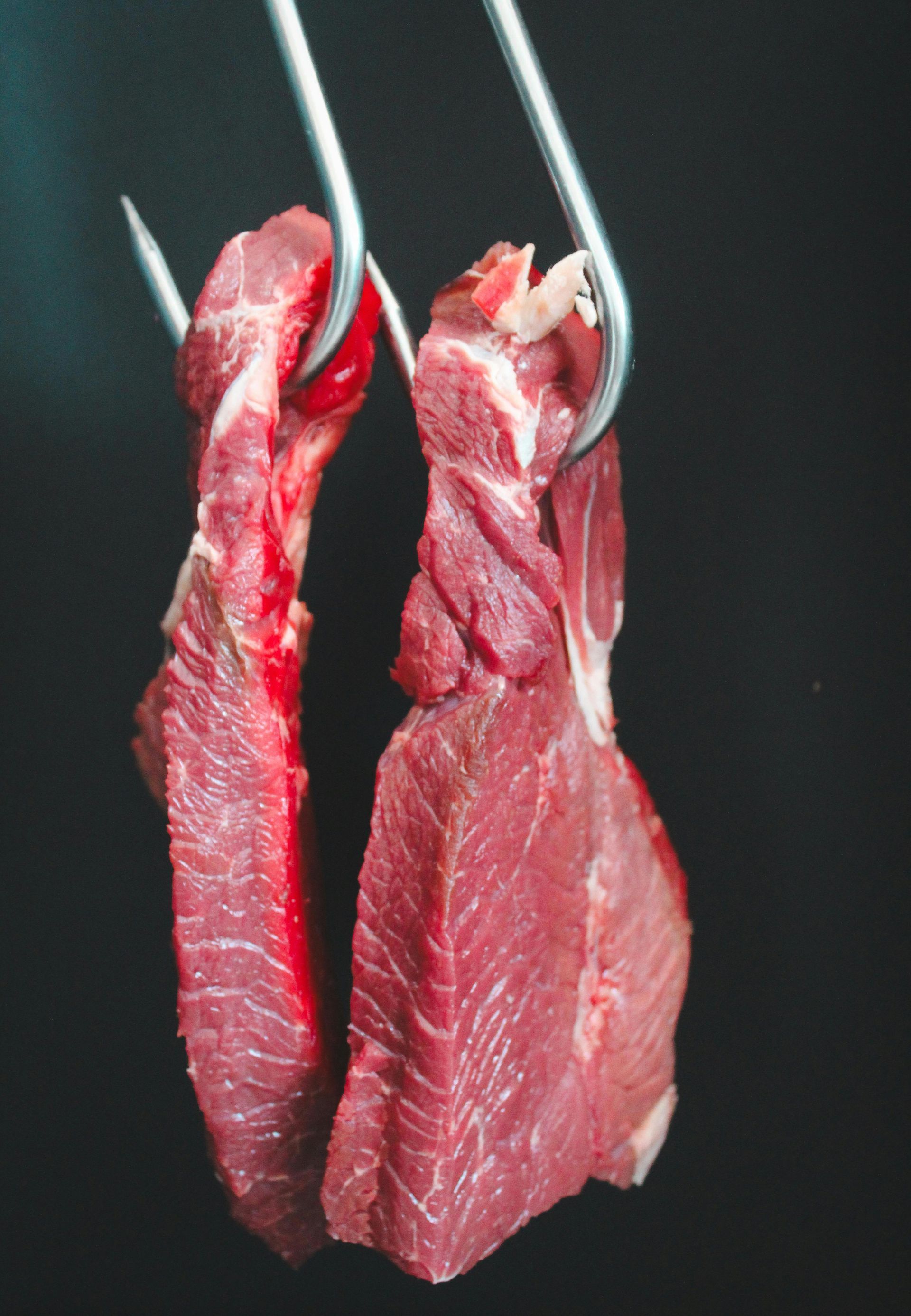 Two raw red beef steaks hanging on metal hooks against a dark background