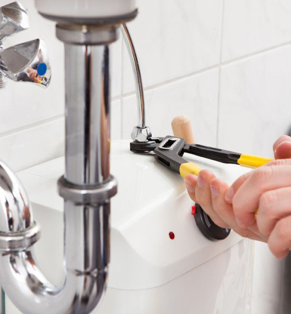 Plumber tightening a nut with a wrench near a chrome pipe and white appliance.