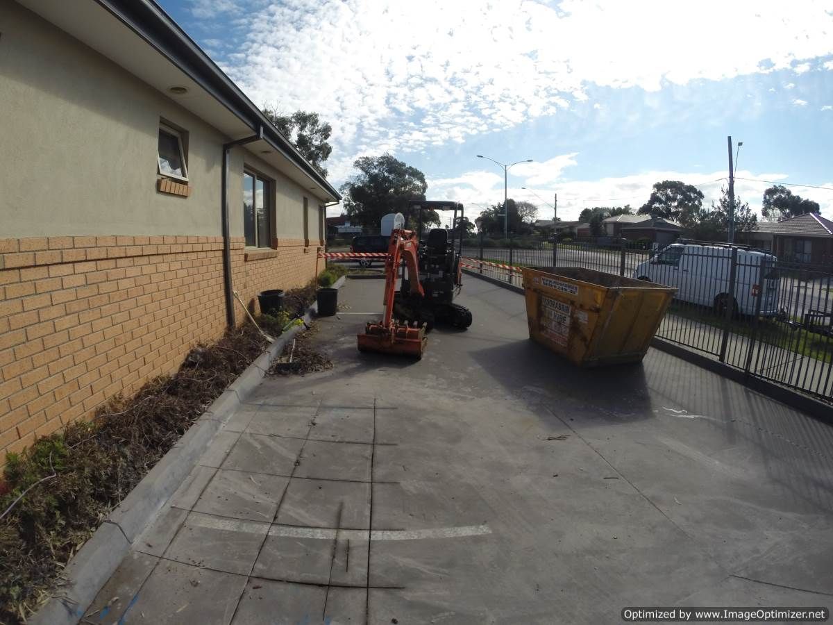 Excavator and skip bin next to a building on a concrete surface, cloudy sky.