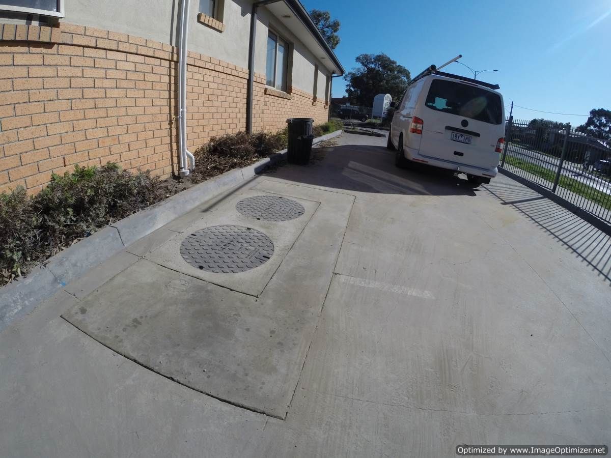 Concrete driveway with manhole covers, trash can, white van parked, beside brick building.