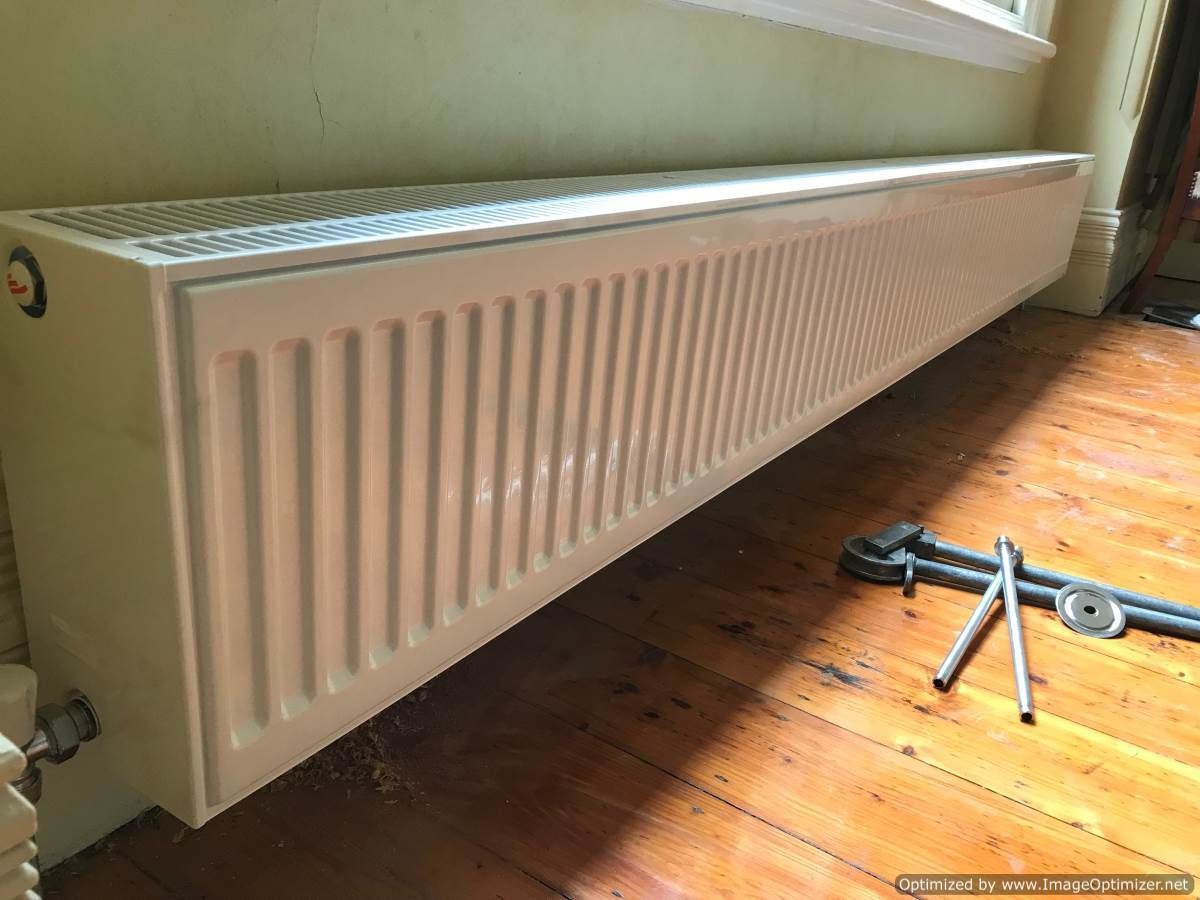 Long, white baseboard radiator against a wall, with tools on the wooden floor.