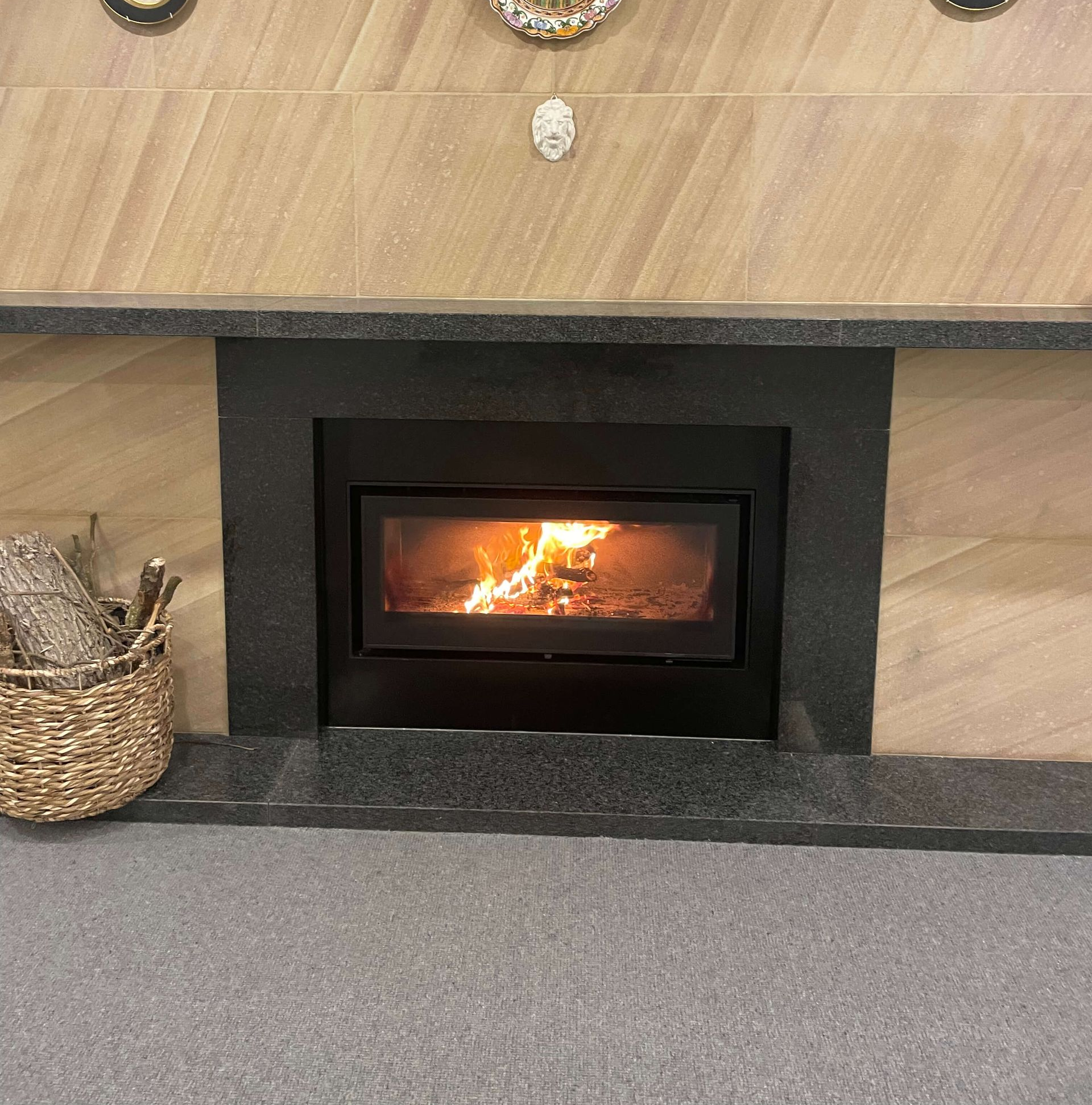 Fireplace with burning logs, black frame, gray hearth and tan wall tile. Basket of firewood at left.