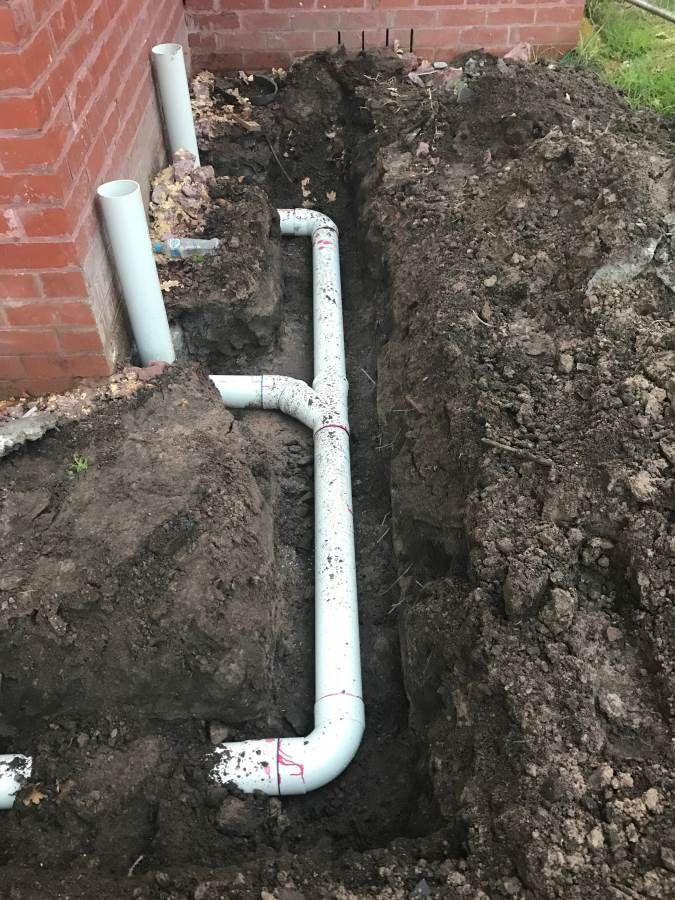 White PVC pipes installed in a trench dug near a red brick building.