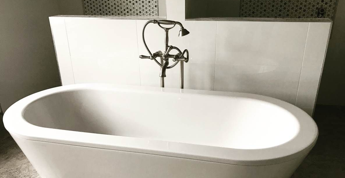 White freestanding bathtub with bronze faucet against a white wall with decorative tile accent.
