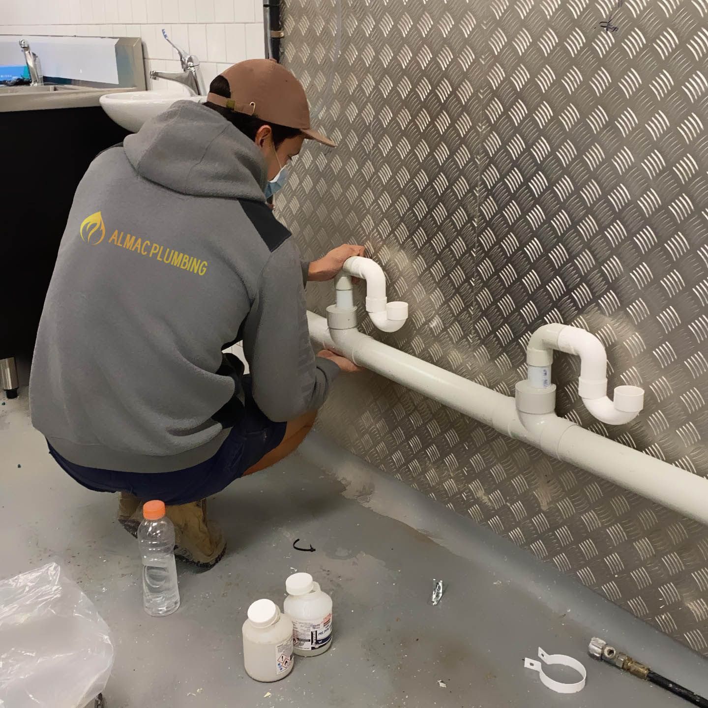 Plumber installing white pipes on a metal wall. He wears a grey hoodie and a baseball cap. Supplies are on the floor.